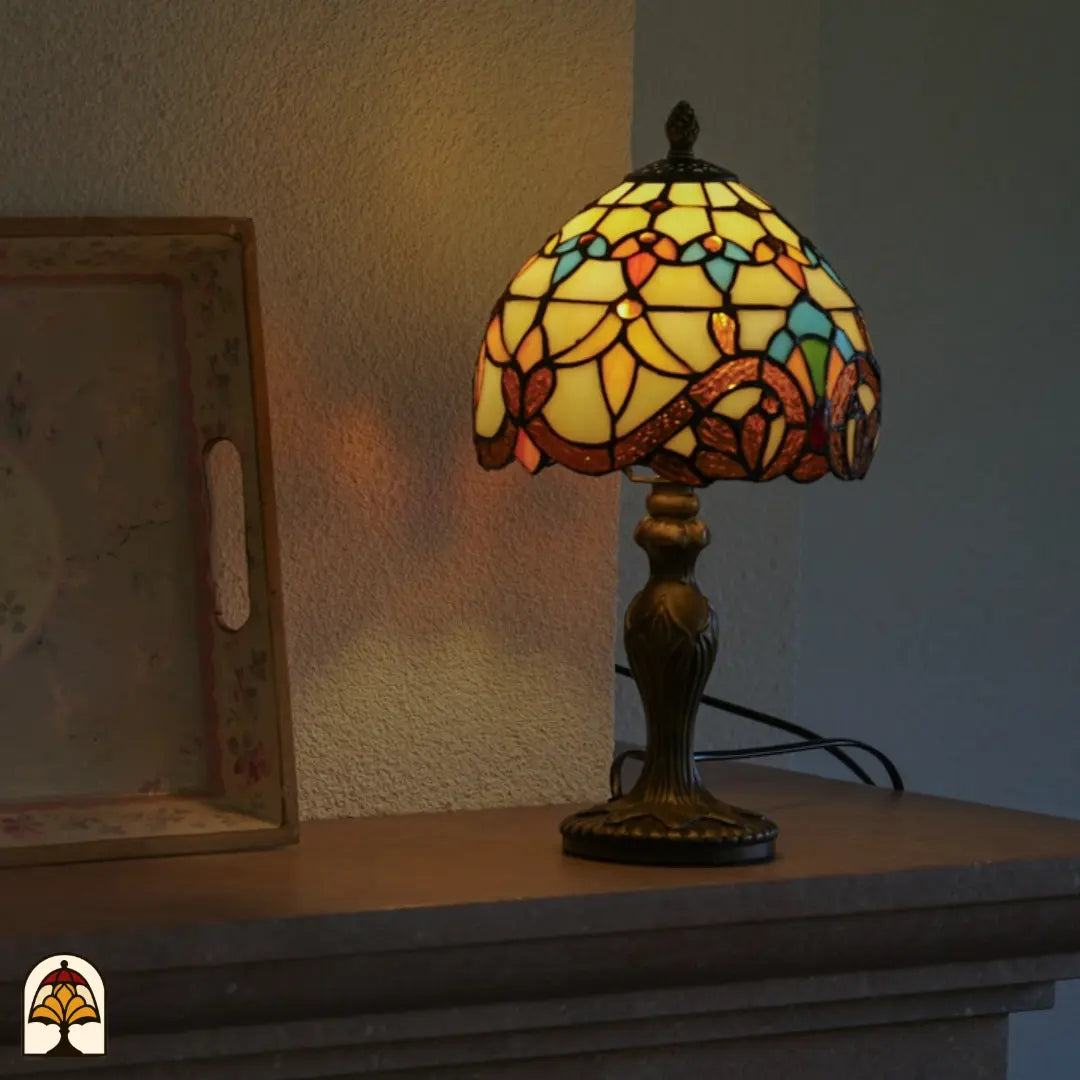Lampe de table Tiffany Baroque allumée sur une cheminée à côté d'un tableau encadré – lueur chaude ambrée et jaune à travers l'abat-jour en dôme baroque