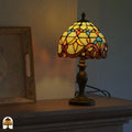 Verlichte Tiffany tafellamp Baroque op schoorsteenmantel naast schilderij in lijst – warme amber en gele gloed door barokke koepelkap