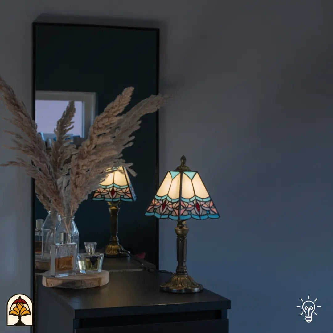 Lifestyle — lampe de table Tiffany allumée sur un buffet noir devant un mur gris, miroir avec reflet, herbes de la pampa et flacons de parfum à côté