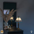 Lifestyle — verlichte Tiffany tafellamp op zwart dressoir voor grijze muur, spiegel met reflectie, pampasgrassen en parfumflesjes ernaast