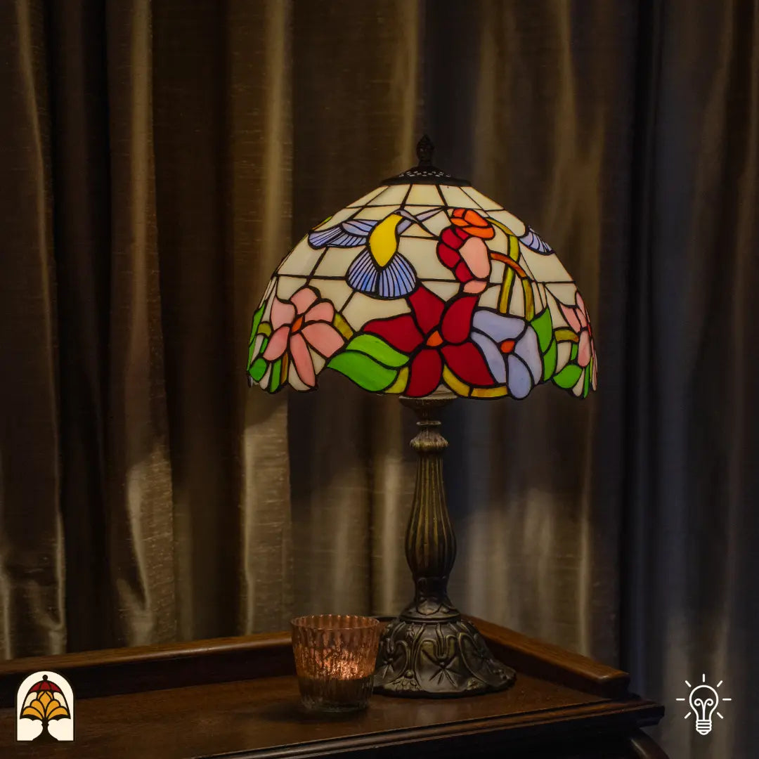 Lampe de table Tiffany Colibrì ⌀30 cm allumée dans un intérieur chaleureux – abat-jour crème avec colibri et fleurs colorées, pied colonne en bronze