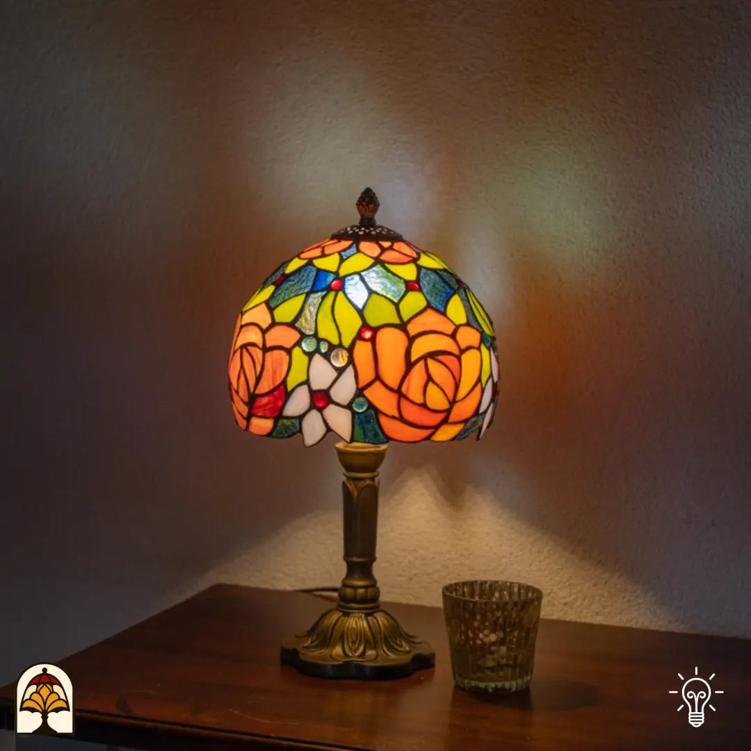 Lampe de table Tiffany Fiore allumée sur une étagère près d'un mur blanc — lumière chaleureuse en arrière-plan, photophore en verre à droite de la lampe
