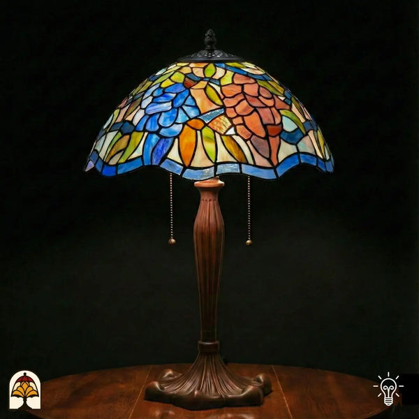 Lampe de table Tiffany Florentia Blu allumée avec large abat-jour en dôme, fleurs bleues et motifs de feuilles orange-rose en vitrail, bordure bleue en bas, pied vase en bronze cannelé avec interrupteurs à chaînette, fond sombre