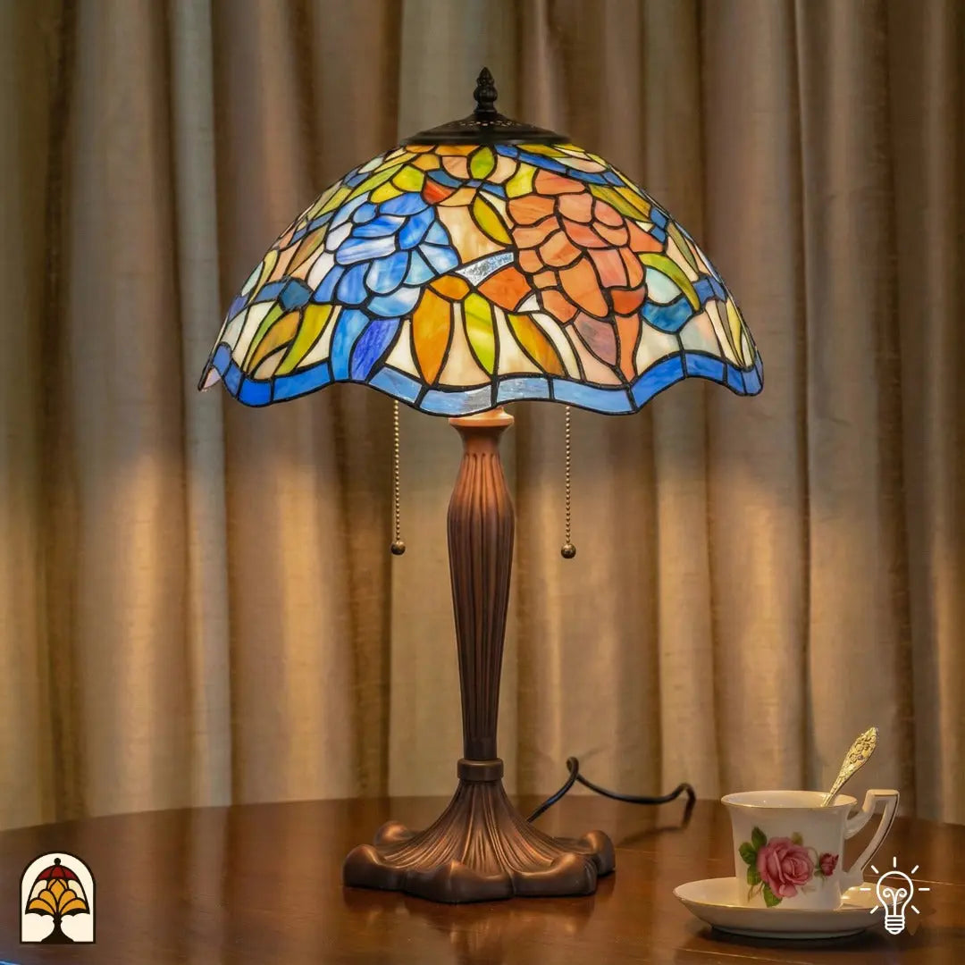 Lampe de table Tiffany Florentia Blu allumée sur une table en bois devant des rideaux, motifs de feuilles bleues et orange illuminés, tasse de thé avec soucoupe à côté