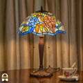Verlichte Tiffany tafellamp Florentia Blu op houten tafel voor gordijnen, blauwe en oranje bladmotieven verlicht, theekopje met schotel ernaast
