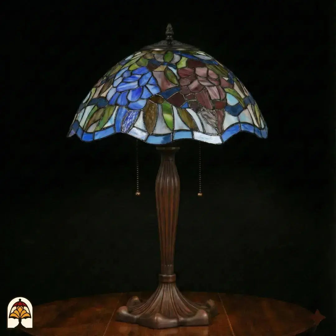 Lampe de table Tiffany Florentia Blu éteinte sur fond sombre, abat-jour avec fleurs bleues et motifs de feuilles violet-brun, pied vase nervuré en bronze avec interrupteurs à chaînette