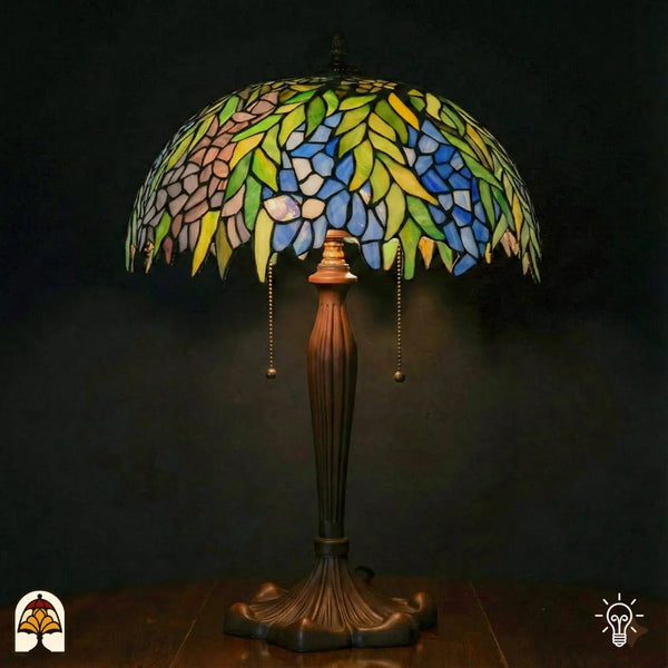 Grande lampe de table Tiffany allumée avec large abat-jour en dôme couvert de feuilles vertes, fleurs bleues et roses/lilas et accents jaunes, pied chandelier en bronze avec interrupteurs à chaînette, fond sombre