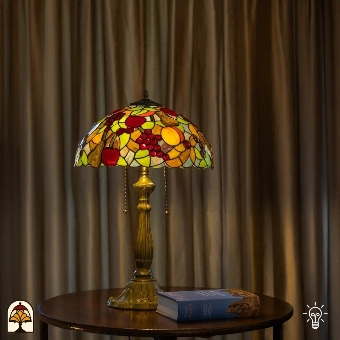 Lampe de table Tiffany Frutta Viva allumée sur une table d'appoint ronde devant des rideaux beiges — livre et objet décoratif à droite de la lampe