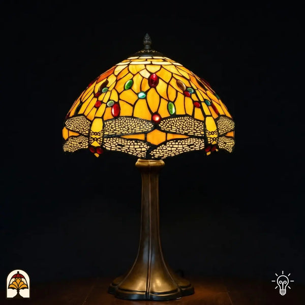 Lampe de table Tiffany Libellula Aurea ⌀30 allumée sur fond sombre, abat-jour en dôme avec panneaux de vitrail jaune doré, frise de libellules en bas avec ailes beiges, cabochons en verre ovales rouges et verts, pied trompette en bronze