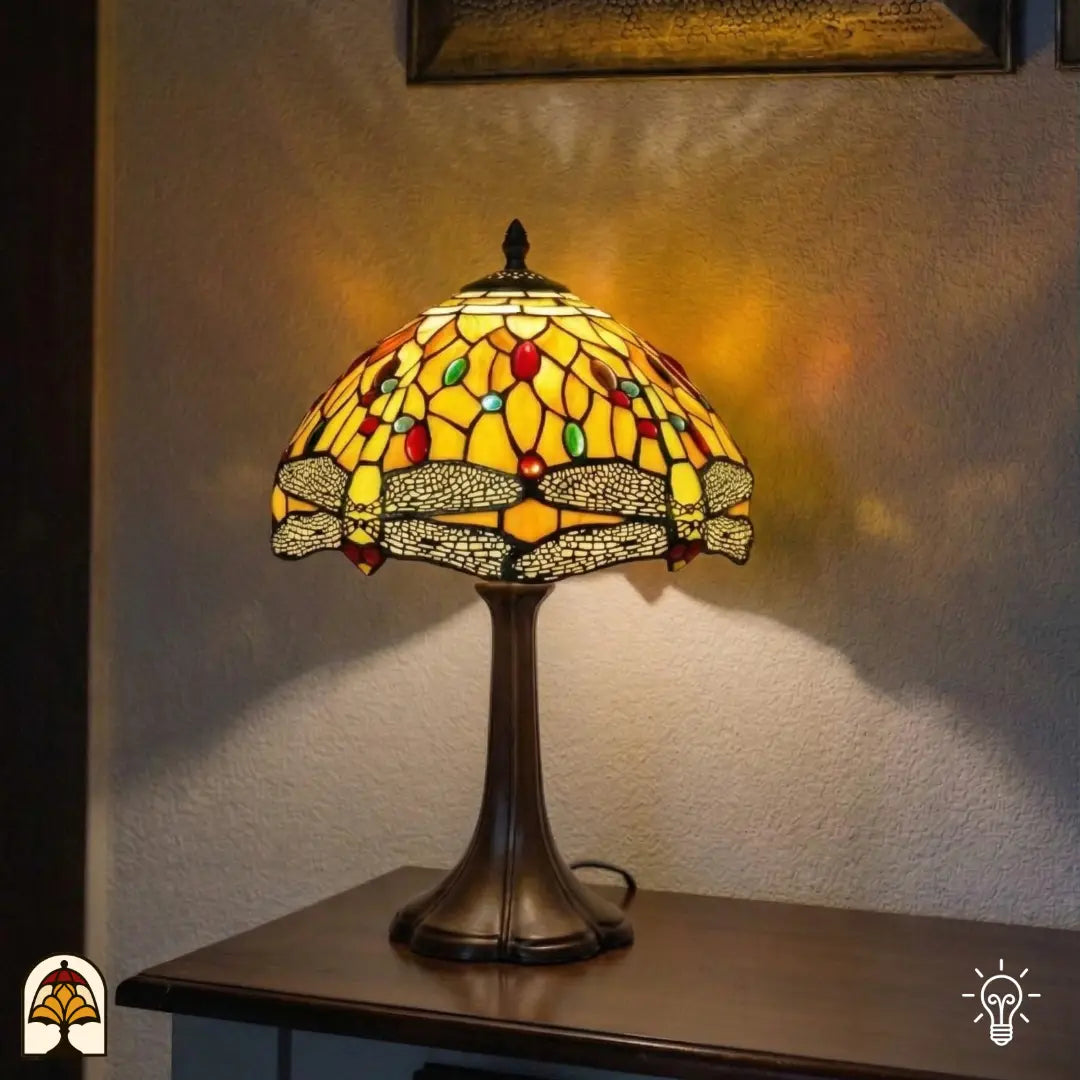 Lampe de table Tiffany Libellula Aurea ⌀30 allumée sur une console dans un angle, lumière chaude jaune-orange sur un mur blanc texturé