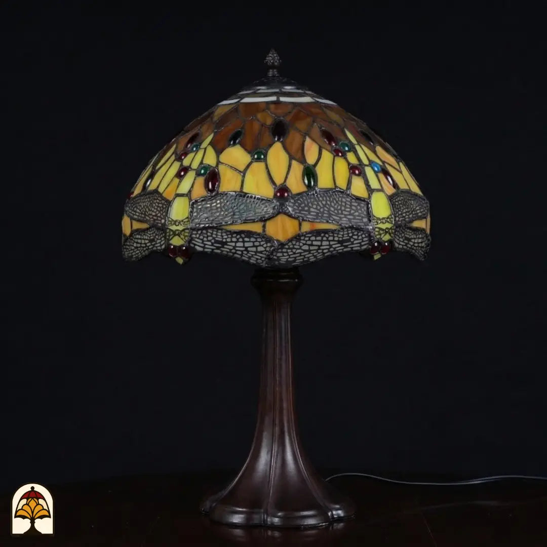 Lampe de table Tiffany Libellula Aurea ⌀30 éteinte sur fond sombre, abat-jour en dôme à panneaux ambré-brun et jaunes, frise de libellules aux ailes grises, cabochons en verre rouges et verts, pied trompette en bronze