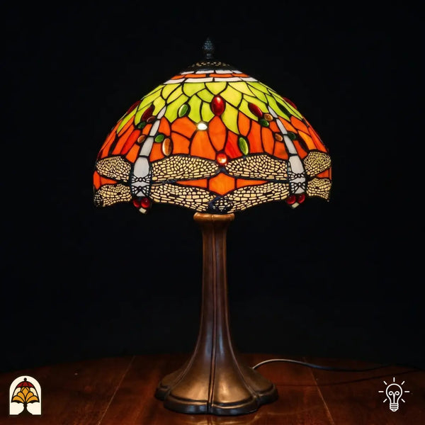 Lampe de table Tiffany Libellula Azul ⌀30 allumée sur fond sombre, abat-jour en dôme avec panneaux de vitrail orange, motifs de feuilles jaune-vert, frise de libellules en bas avec ailes beiges, cabochons en verre ovales rouges et verts, pied trompette en bronze