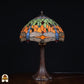 Onverlichte Tiffany tafellamp Libellula Azul ⌀30 op donkere achtergrond, koepelvormige kap met oranje panelen, groene bladmotieven, blauwe libellenmotieven en blauwe glasaccenten, rode en groene ovale juwelen, bronzen trompetvoet