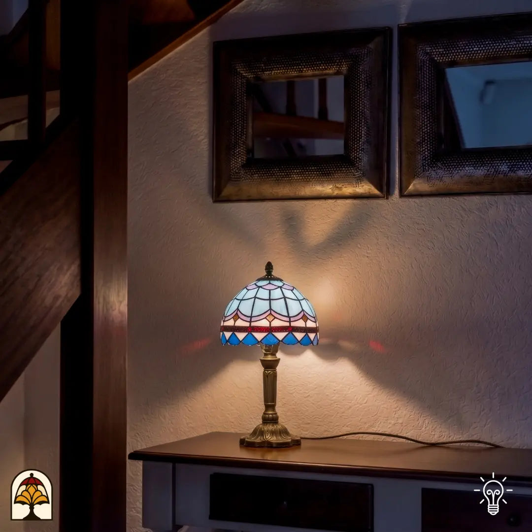 Une petite lampe de table Tiffany pleine d'atmosphère avec un abat-jour à motif d'écailles bleu et blanc et une bordure décorative foncée. La lampe repose sur un pied élégant couleur bronze posé sur un buffet en bois. Devant un mur blanc avec des miroirs décoratifs et un escalier en bois, la lampe diffuse une lumière douce et chaleureuse qui se reflète subtilement sur le mur.