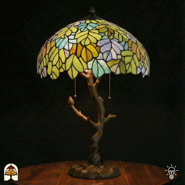 Lampe de table Tiffany Sylva Verde allumée avec large abat-jour en dôme couvert de motifs de feuilles jaune-vert, jaune-orange et bleu clair en vitrail, pied en forme de tronc d'arbre en bronze patiné avec ramifications et interrupteurs à chaînette, fond sombre