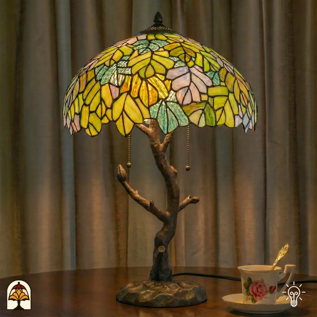 Lampe de table Tiffany Sylva Verde allumée sur une table en bois devant des rideaux, motifs de feuilles jaune-vert et orange illuminés, tasse de thé avec soucoupe à côté
