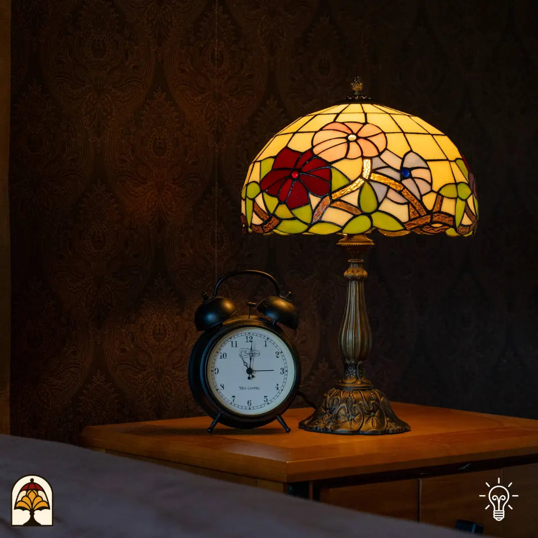 Lampe de table Tiffany Aurora Bloom allumée sur une table de chevet en bois — papier peint sombre au mur, réveil ancien à gauche de la lampe