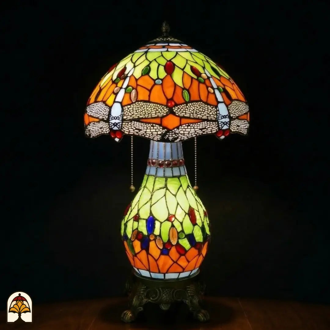 Lampe de table Tiffany Libellula Lumina ⌀30 allumée sur fond sombre, abat-jour en dôme avec panneaux orange et jaune-vert, frise de libellules, pied sphérique en vitrail avec panneaux jaune-vert et orange et cabochons en verre colorés, pied en bronze à pattes de lion, interrupteurs à chaînette