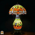 Verlichte Tiffany tafellamp Libellula Lumina ⌀30 op donkere achtergrond, koepelvormige kap met oranje en geel-groene panelen, libellenfries, bolvormige glas-in-lood voet met geel-groene en oranje panelen en gekleurde juwelen, bronzen leeuwenpootvoet, trekschakelaars