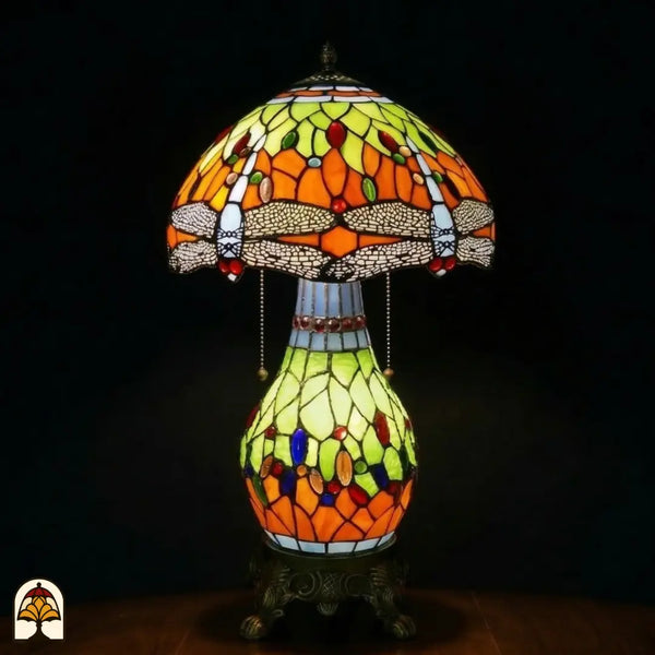 Lampe de table Tiffany Libellula Lumina ⌀30 allumée sur fond sombre, abat-jour en dôme avec panneaux orange et jaune-vert, frise de libellules, pied sphérique en vitrail avec panneaux jaune-vert et orange et cabochons en verre colorés, pied en bronze à pattes de lion, interrupteurs à chaînette
