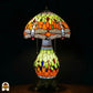 Verlichte Tiffany tafellamp Libellula Lumina ⌀30 op donkere achtergrond, koepelvormige kap met oranje en geel-groene panelen, libellenfries, bolvormige glas-in-lood voet met geel-groene en oranje panelen en gekleurde juwelen, bronzen leeuwenpootvoet, trekschakelaars