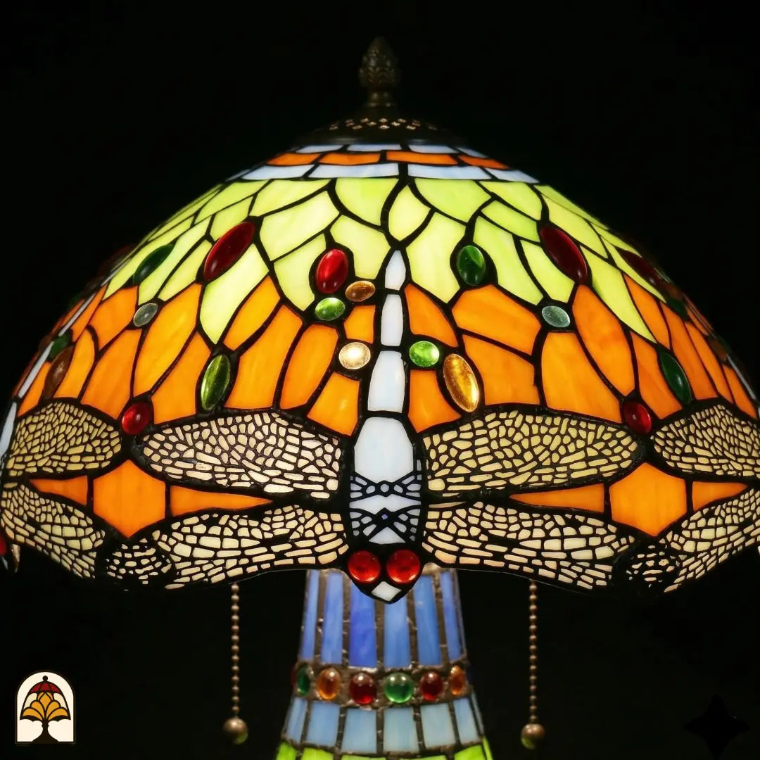 Gros plan abat-jour Libellula Lumina ⌀30 allumé, panneaux de vitrail orange et jaune-vert avec cabochons rouges, verts et ambrés, frise de libellules aux ailes blanches, dessus du pied sphérique en vitrail visible