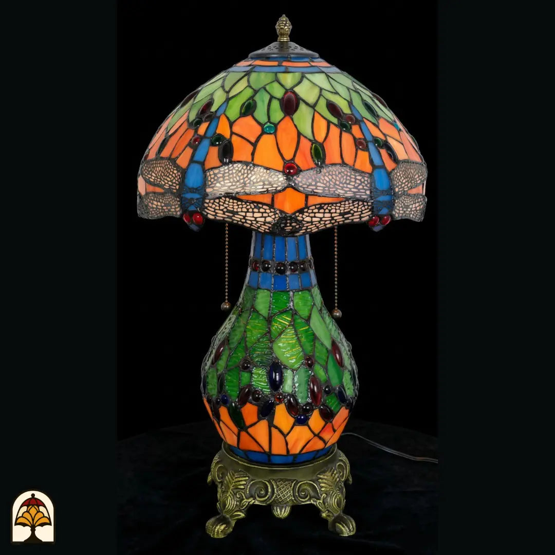 Lampe de table Tiffany Libellula Lumina ⌀30 éteinte sur fond sombre, abat-jour à panneaux verts et orange, frise de libellules bleue, cabochons en verre colorés, pied boule en vitrail vert, interrupteurs à chaînette, base à pattes de lion en bronze