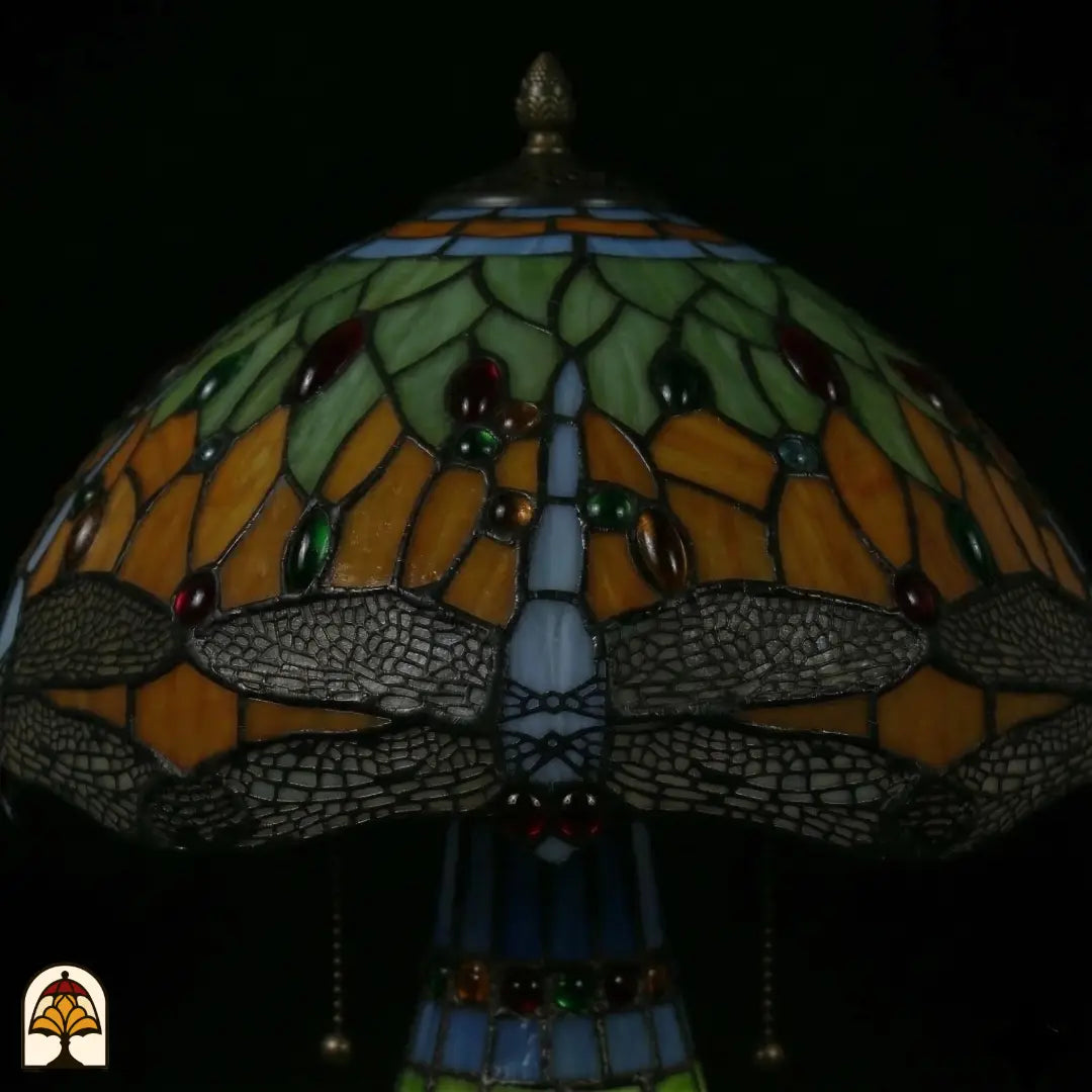 Gros plan abat-jour Libellula Lumina ⌀30 éteint, panneaux de vitrail verts et orange avec cabochons rouges, verts et ambrés, frise de libellules bleue le long du bord