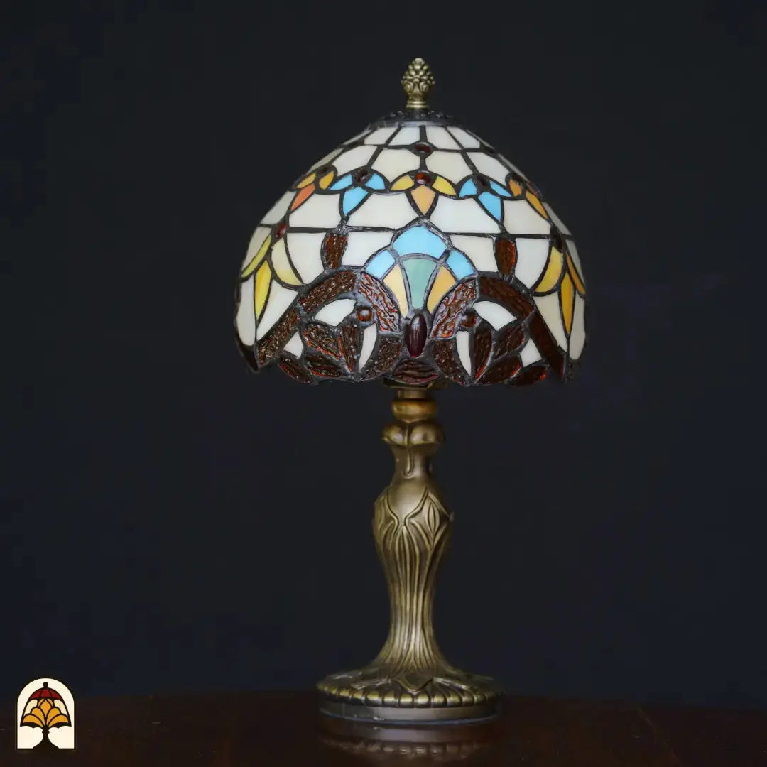 Lampe de table Tiffany Baroque éteinte sur fond noir – abat-jour dôme avec formes de feuilles baroques crème, bleu clair, jaunes et brunes, pierre précieuse rouge