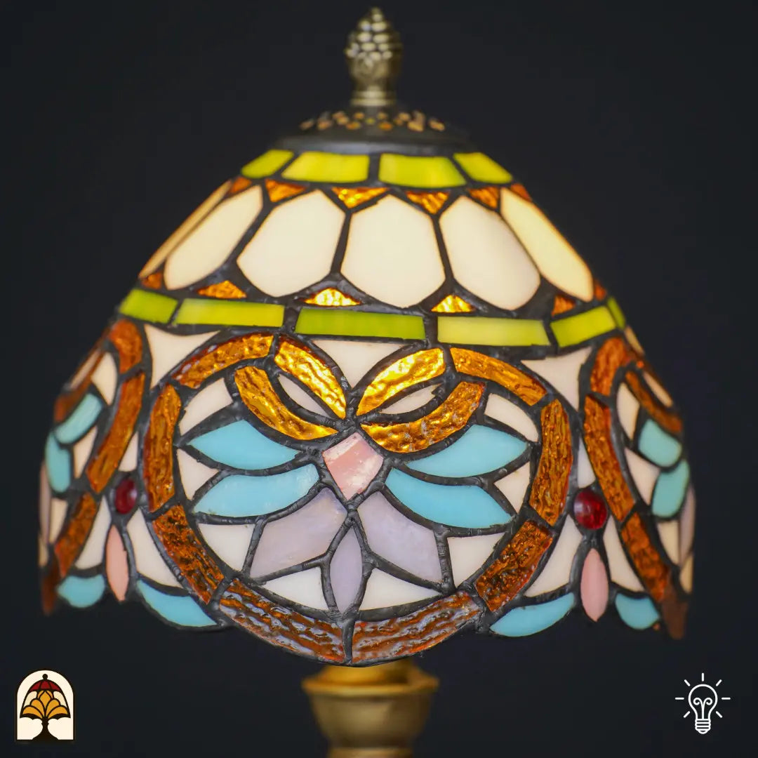 Gros plan abat-jour en dôme allumé lampe de table Tiffany Lucette – formes de feuilles ambre doré avec pétales bleu clair et roses, bandeau décoratif vert et cabochons en verre rouges