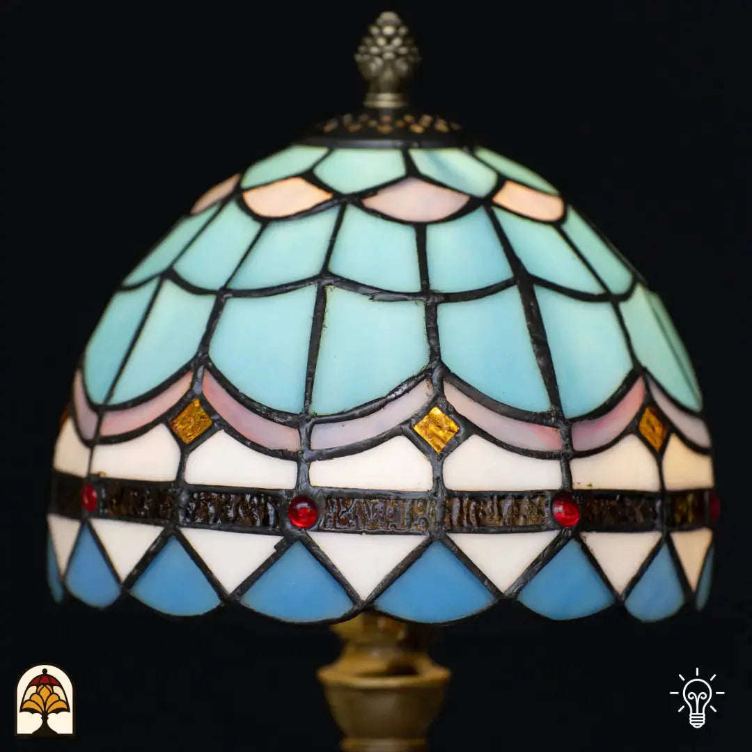 Gros plan abat-jour en dôme allumé lampe de table Tiffany Sapphire – écailles de poisson bleu clair et roses en vitrail, cabochons en verre dorés en forme de diamant et cabochons rouges sur bandeau décoratif