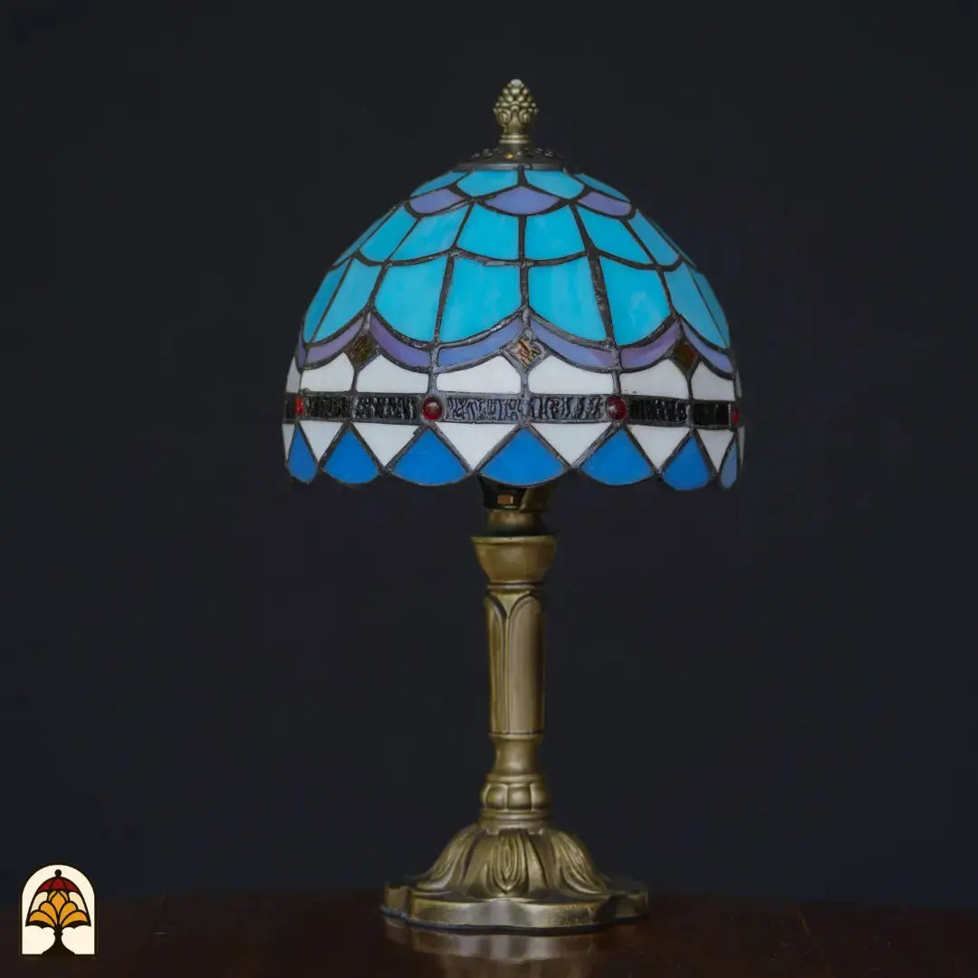 Lampe de table Tiffany Sapphire éteinte sur fond sombre – abat-jour dôme bleu clair et blanc à motif d'écailles de poisson, accents violets, cabochons en verre rouges, pied colonne en bronze