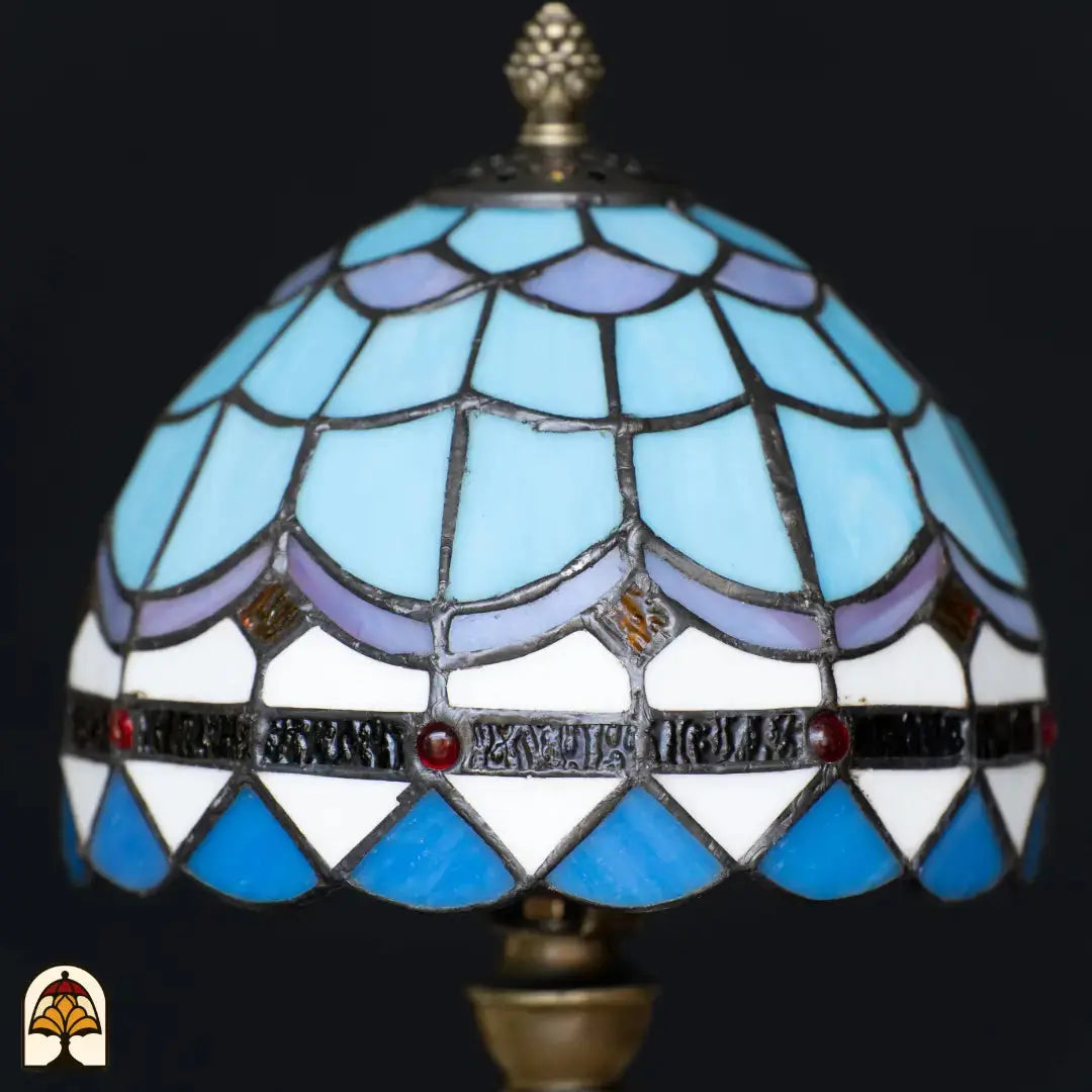 Gros plan abat-jour dôme éteint lampe de table Tiffany Sapphire – motif d'écailles de poisson bleu et blanc avec ligne ondulée violette, losanges dorés et cabochons rouges sur le bandeau décoratif