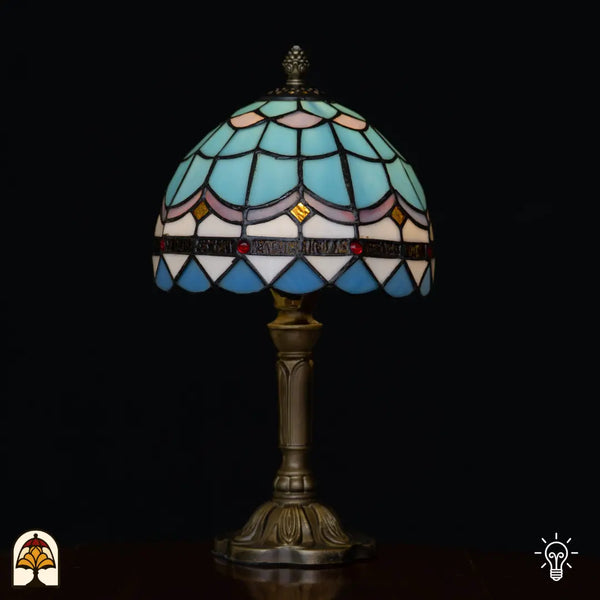 Lampe de table Tiffany Sapphire ⌀20 cm allumée sur fond noir – abat-jour en dôme bleu clair à motif d'écailles de poisson, bandeau décoratif rose, cabochons en verre rouges et pied en bronze