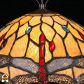 Close-up verlichte kap Tiffany hanglamp Libelle, geel-amber glas-in-lood met rode druppels en blauwe accenten, libellenmotieven langs de rand
