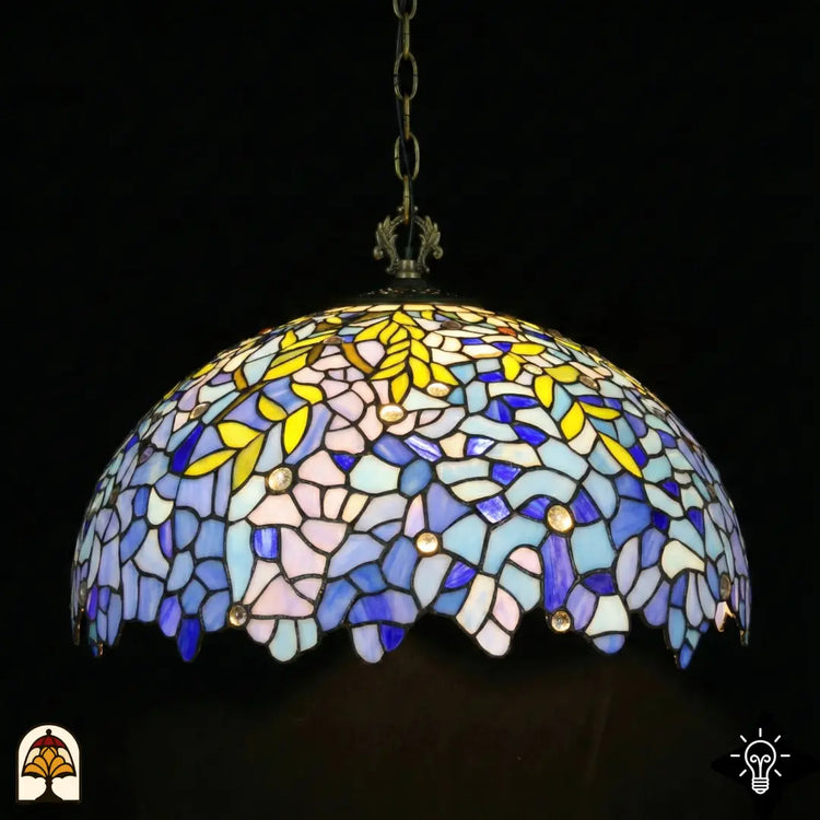 Onverlichte Tiffany hanglamp Drizzle Bleu ⌀50 op donkere achtergrond, koepelkap met lila-blauwe en turquoise glas-in-lood panelen, gele bladmotieven, ronde kristalaccenten, geschulpte onderrand, bronzen ketting