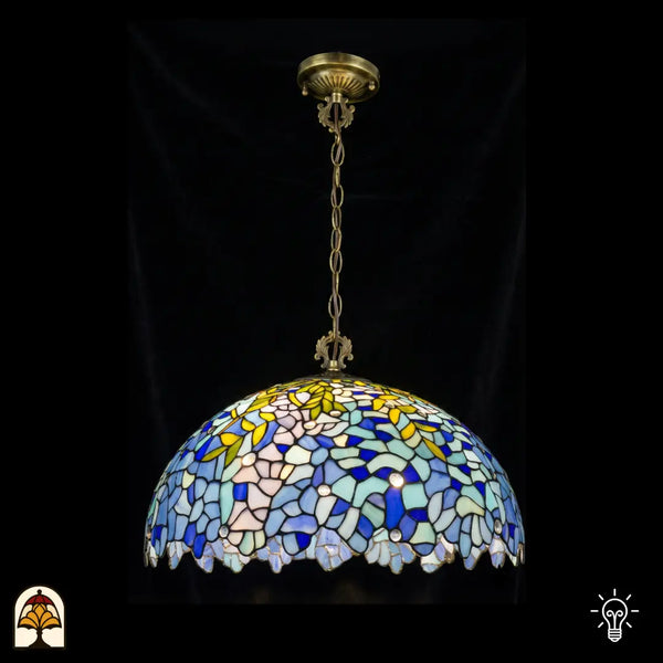Suspension Tiffany Drizzle Bleu ⌀50 allumée sur fond sombre, grand abat-jour en dôme à motifs floraux en vitrail bleu et turquoise, accents de feuilles jaunes, bordure festonnée, chaîne en bronze et rosace de plafond