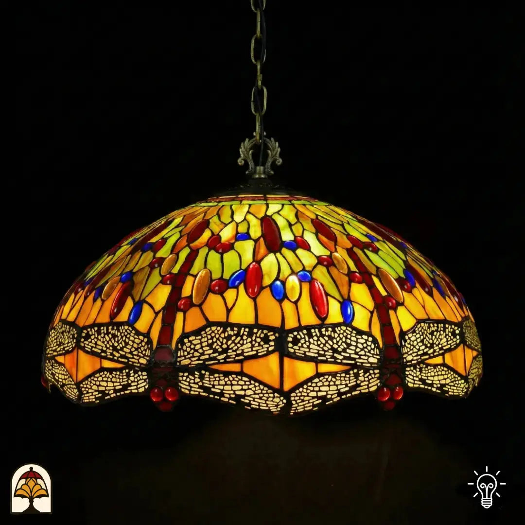 Suspension Tiffany Libellula Aurelia ⌀50 allumée, abat-jour en dôme avec panneaux de vitrail jaunes et verts, cabochons en verre rouges et bleus, frise de libellules le long du bord, chaîne en bronze