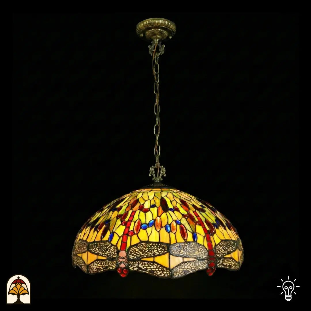 Suspension Tiffany Libellula Aurelia ⌀50 éteinte sur fond sombre, grand abat-jour en dôme à panneaux de vitrail jaunes, frise de libellules en bas, cabochons en verre rouges et bleus, chaîne en bronze et rosace de plafond