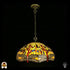 Onverlichte Tiffany hanglamp Libellula Aurelia ⌀50 op donkere achtergrond, grote koepelvormige kap met gele glas-in-lood panelen, libellenfries onderaan, rode en blauwe juwelen, bronzen ketting en plafondrozet