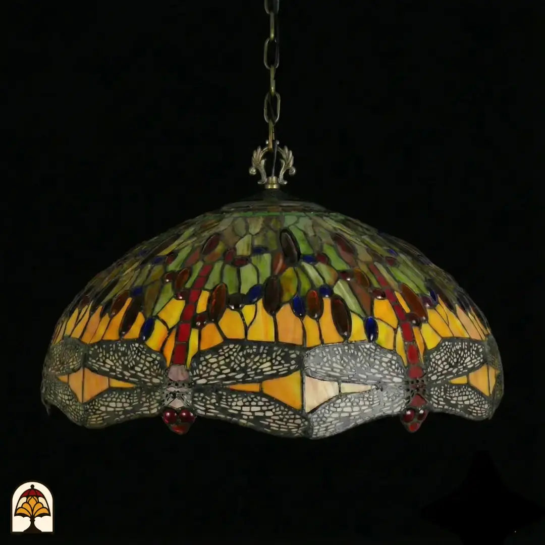 Suspension Tiffany Libellula Aurelia ⌀50 éteinte sur fond sombre, abat-jour à panneaux jaunes et verts, frise de libellules grise avec cabochons en verre rouges le long du bord, chaîne en bronze