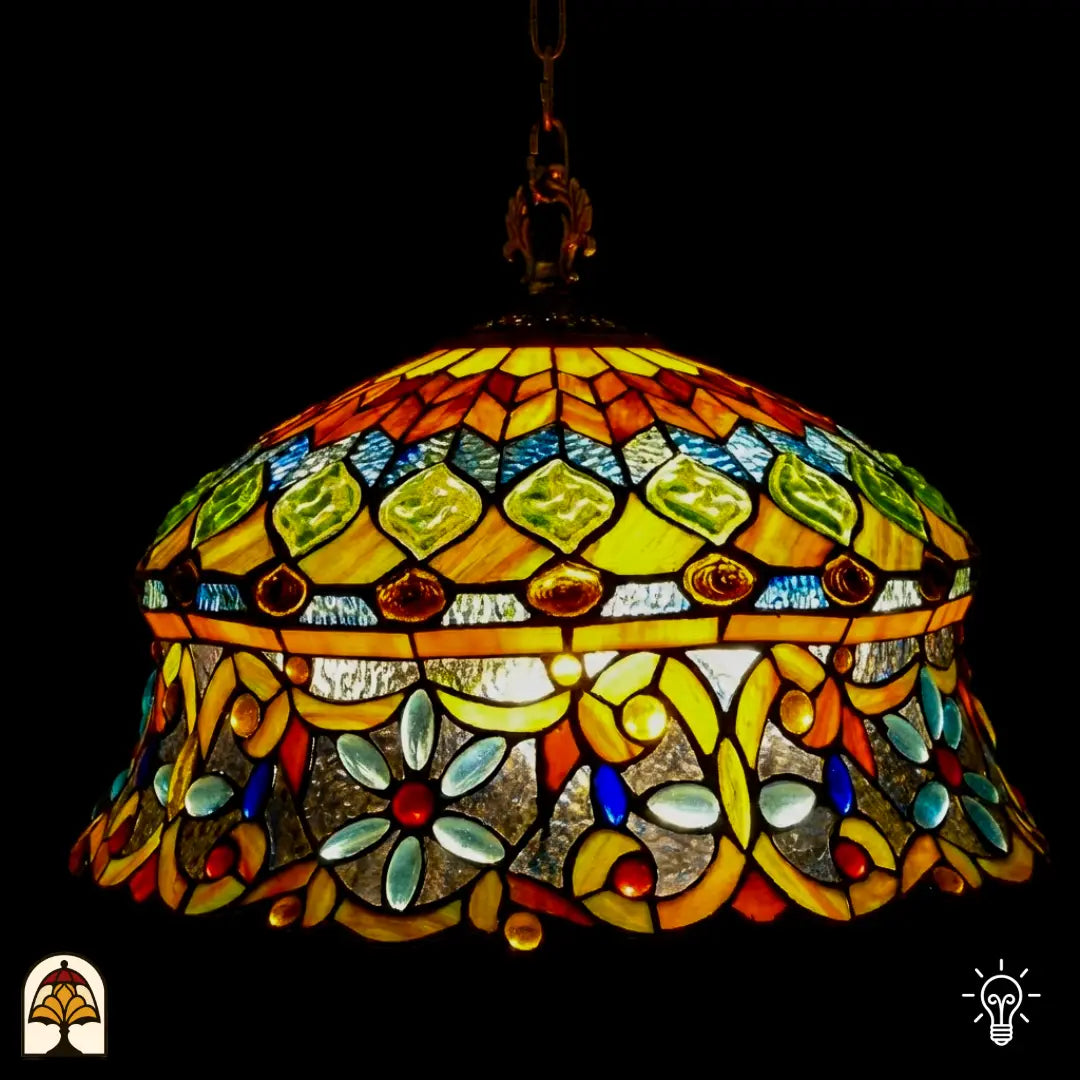 Suspension Tiffany allumée en gros plan, abat-jour en dôme avec zone supérieure ambre-jaune à cabochons en verre verts et cabochons ovales brun-rouge, zone inférieure avec arabesques jaunes, cabochons en forme de fleur bleu clair et cabochons ronds rouges sur verre transparent