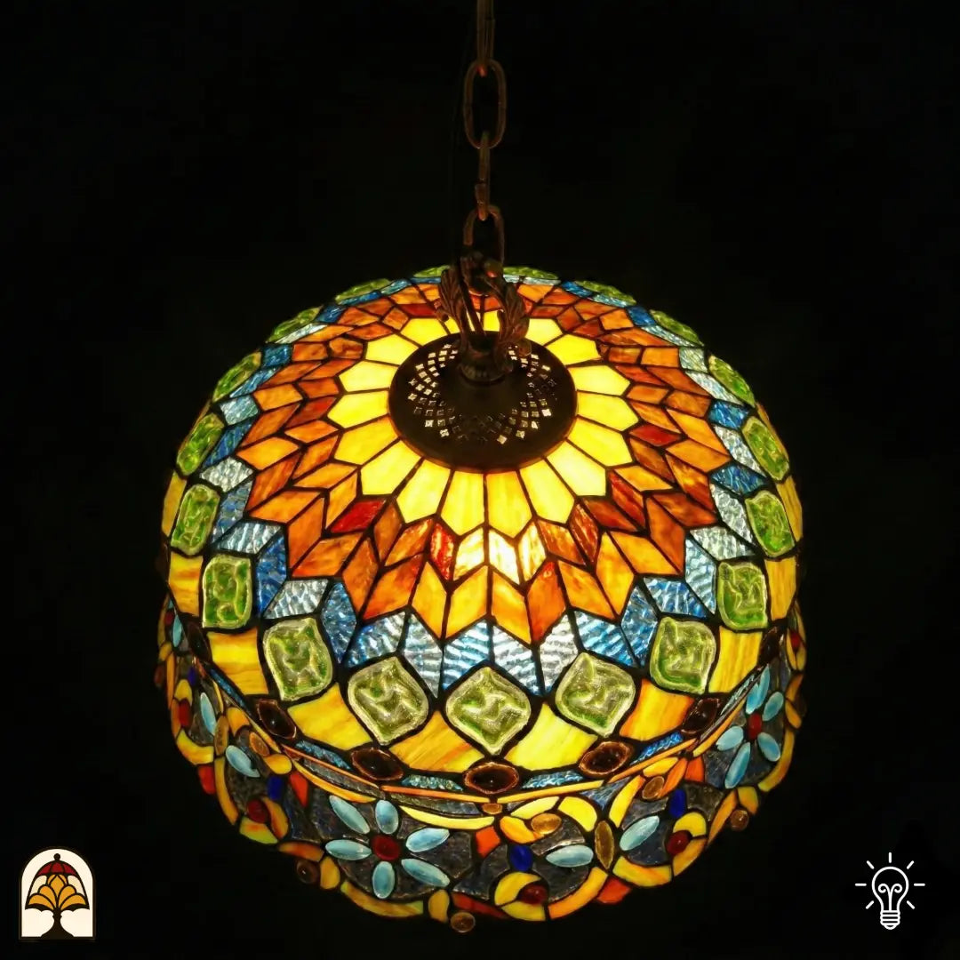 Vue de dessus de suspension lampe Tiffany allumée, motif rayonnant avec surfaces ambrées, cabochons en verre verts et cases en mosaïque bleues, plaque d'aération en bronze visible au centre