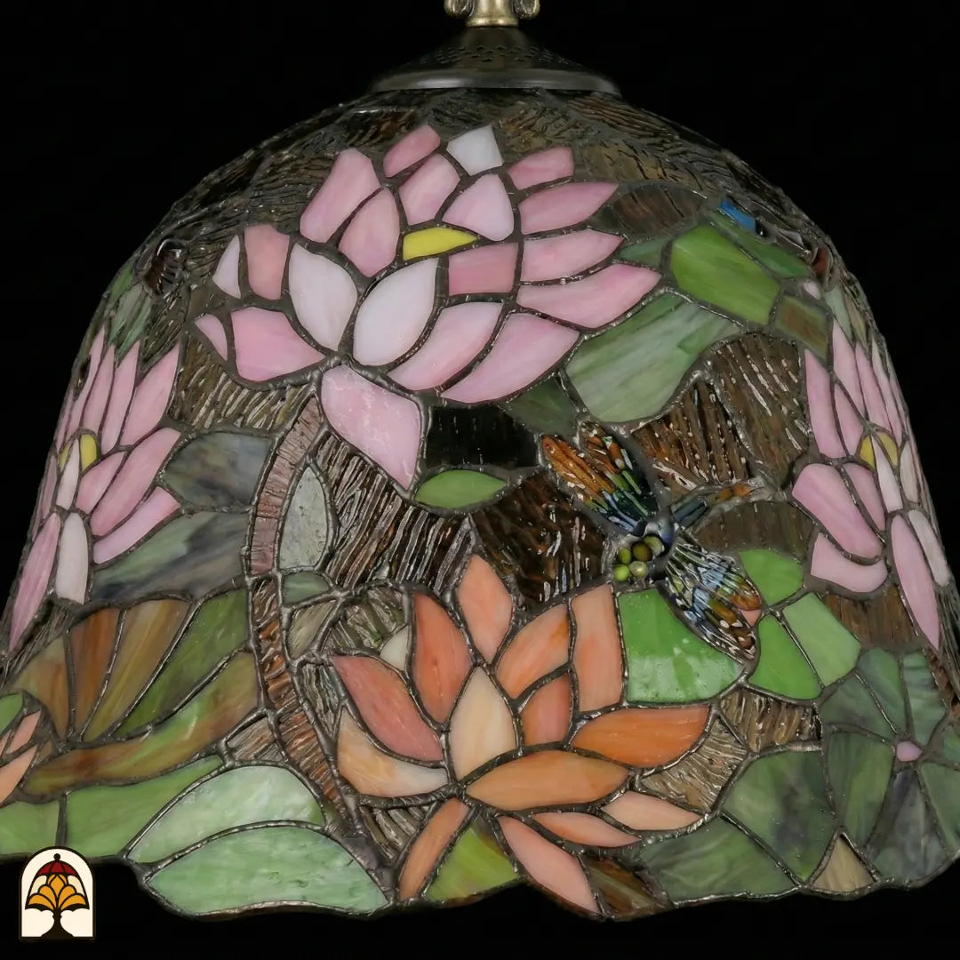 Gros plan de lampe Tiffany, abat-jour en forme de cloche éteint avec fleur de lotus rose au centre, fleurs de lotus orange en bas, feuilles de nénuphar vertes et une libellule brun-orange avec détails bleus