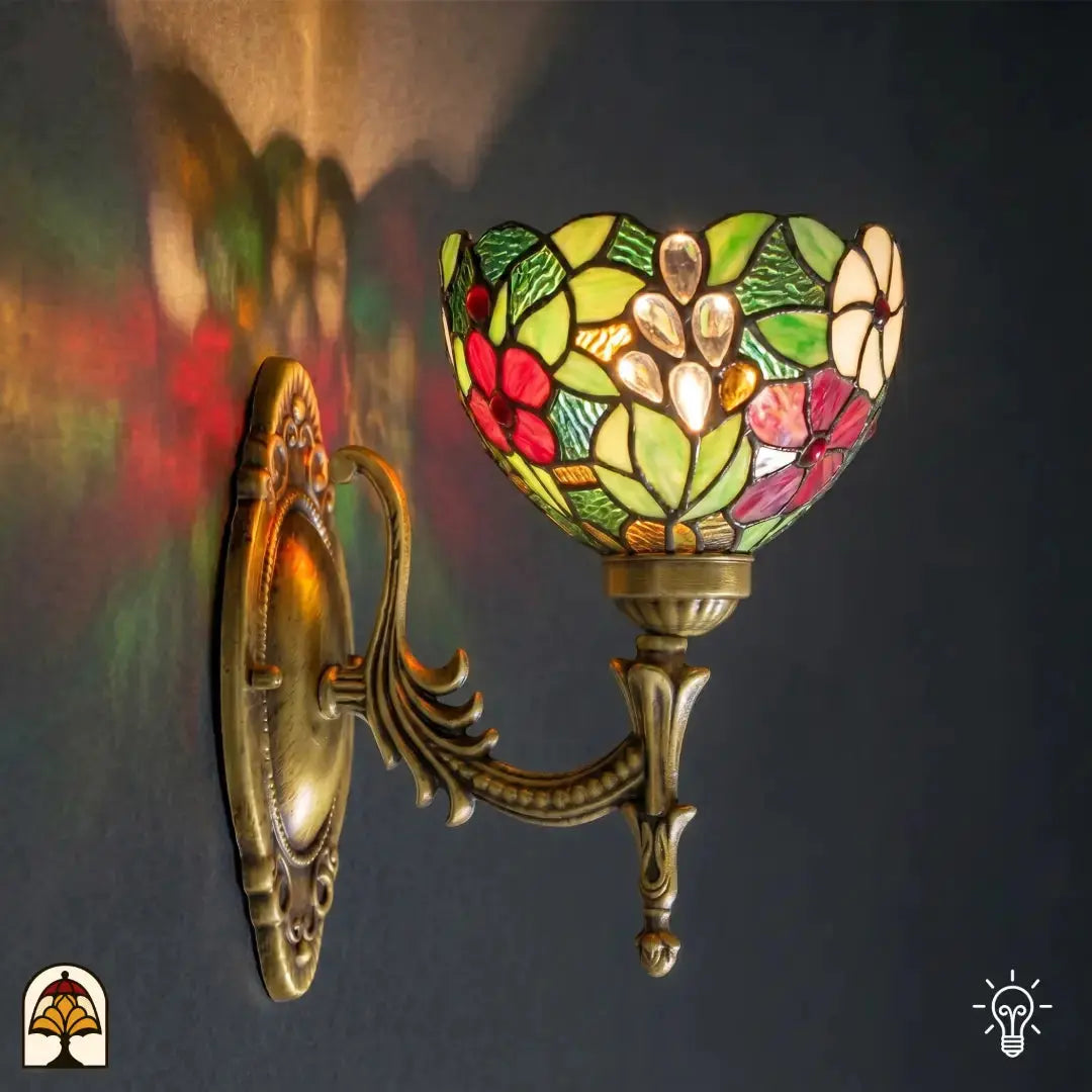 Applique murale Tiffany allumée au mur, abat-jour en vitrail en forme de coupe avec fleurs rouges et roses, feuilles vertes et cabochons en verre transparents ; support mural en bronze orné avec platine ovale et rinceaux d'acanthe ; reflets de lumière colorés sur le mur