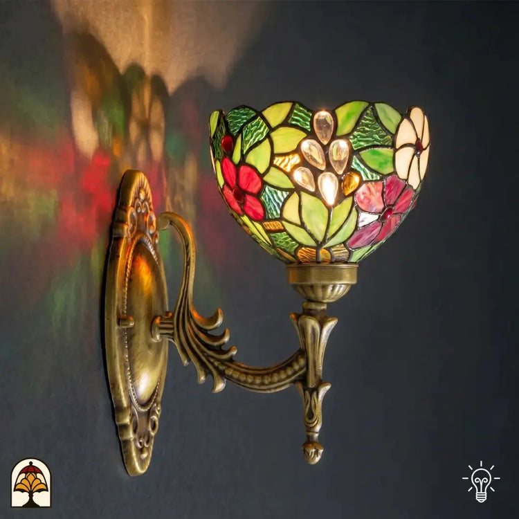 Tiffany lamp wandlamp verlicht aan muur, komvormige glas-in-lood kap met rode en roze bloemen, groene bladeren en transparante glaskralen; sierlijke bronzen muurbeugel met ovale wandplaat en acanthusrolwerk; kleurrijke lichtreflecties op de muur