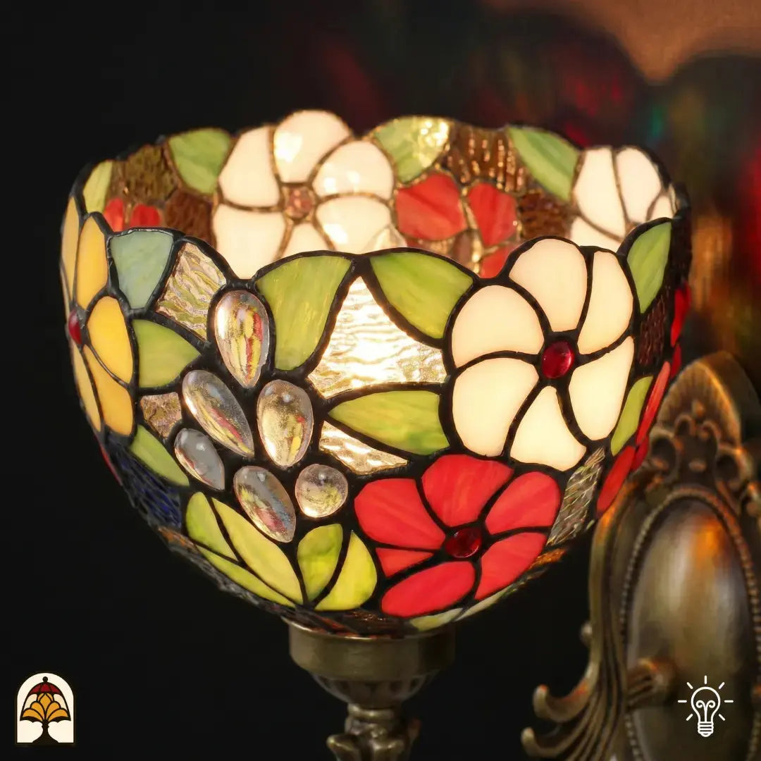 Gros plan d'applique murale Tiffany, abat-jour en vitrail allumé, abat-jour en forme de coupe avec fleurs blanches, fleurs rouges, feuilles vertes et cabochons en verre transparents en forme de goutte sur fond brun ambré, douille en bronze visible