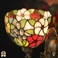Detailopname van tiffany lamp wandlamp glas-in-lood kap verlicht, komvormige kap met witte bloemen, rode bloemen, groene bladeren en druppelvormige transparante glaskralen op bruinamberen achtergrond, bronzen fitting zichtbaar