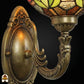 Close-up van bronzen wandbeugel met ovale reliëfplaat en gebogen arm, bovenkant van de florale glas-in-lood kap met groen en amber zichtbaar