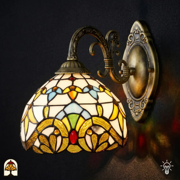 Applique murale Tiffany Barok allumée vue de face avec abat-jour en vitrail jaune doré, accents bleus et verts, sur bras courbé en bronze avec platine murale ovale